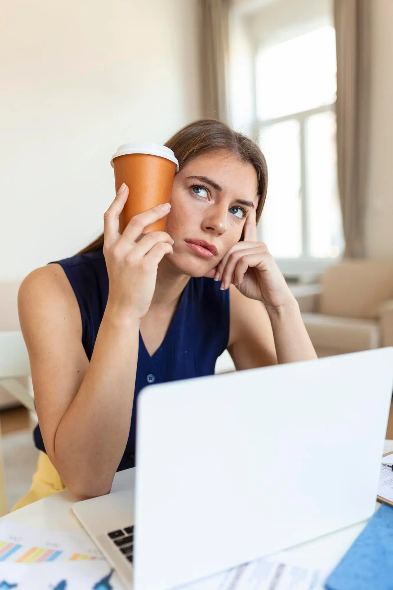 Jeune femme fatiguée dans un bureau, le regard détourné, tenant une tasse de café près de sa tête, avec un premier plan flou.